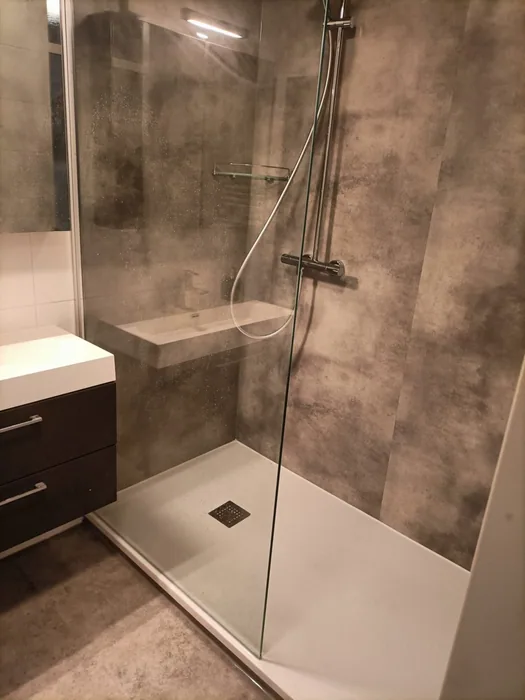 Douche met bruingrijze betonlook panelen, witte douchevloer, glazen wand en wastafelmeubel