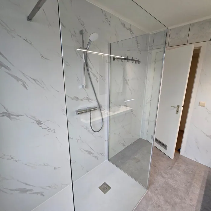 Ruime inloopdouche met glazen wand, thermostaatkraan en witte marmerlook wandpanelen, deur naar de gang open