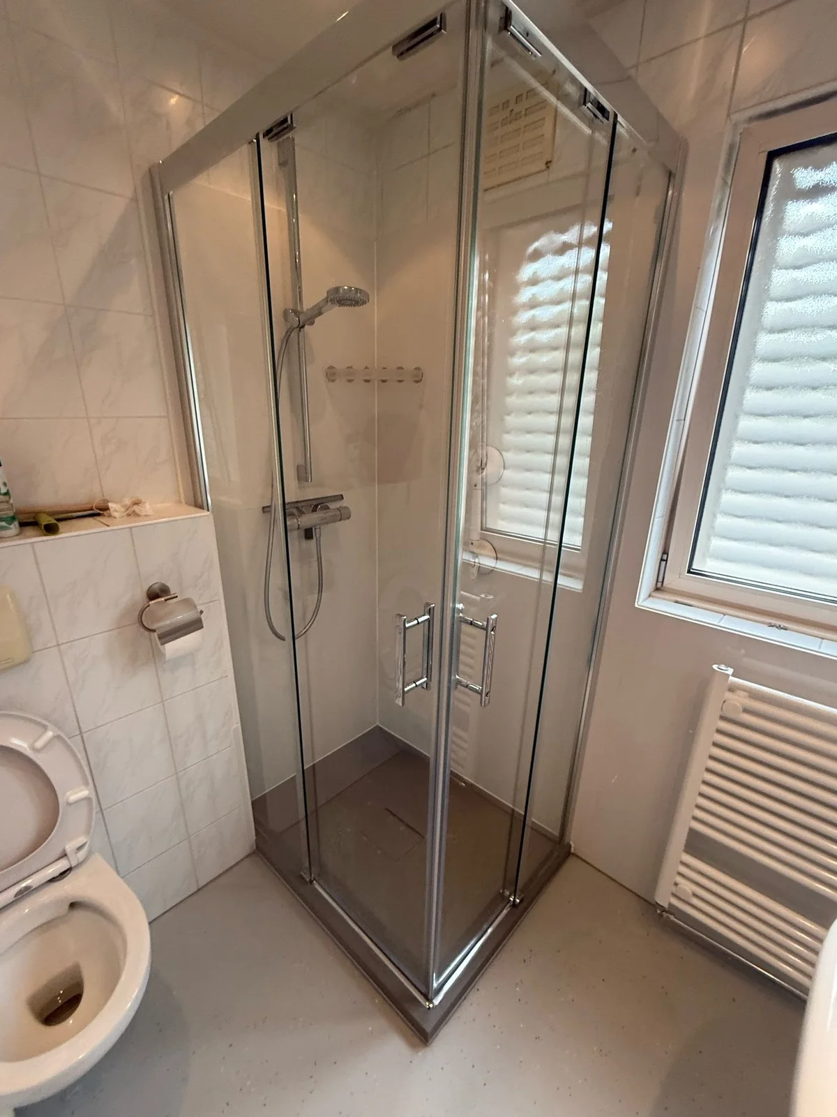 Voor: Vierkante glazen douchecabine naast staand toilet en handdoekradiator, nog zonder steunbeugel