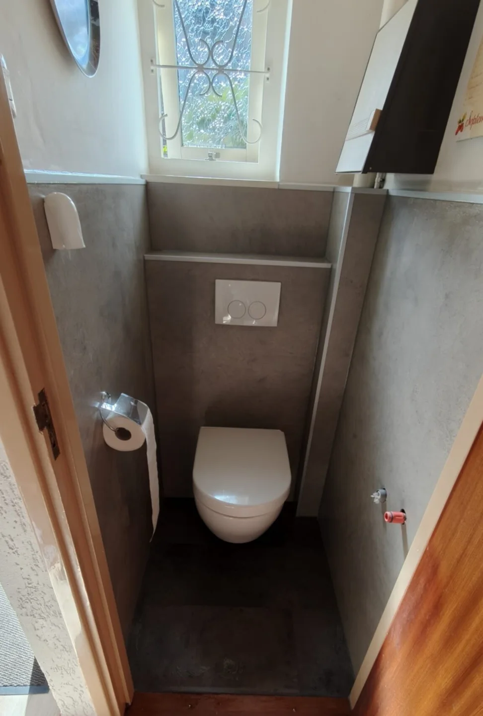 Na: Gerenoveerd toilet met hangtoilet, grijze betonlook wandpanelen, voorzetwand met bedieningsplaat en raampje met sierrooster