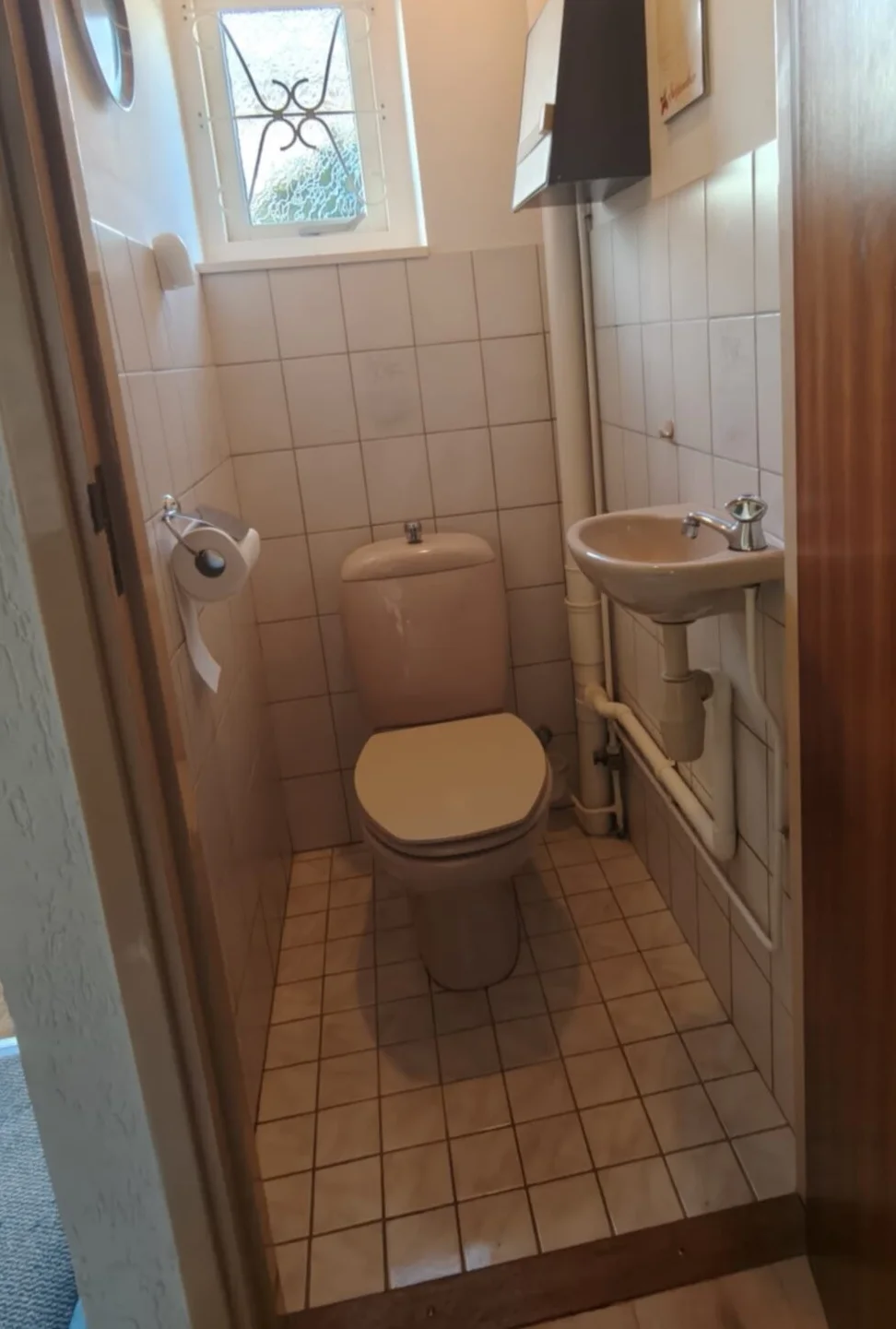 Voor: Oud toilet met beige staand closet, klein fonteintje, witte tegels en raampje met sierrooster